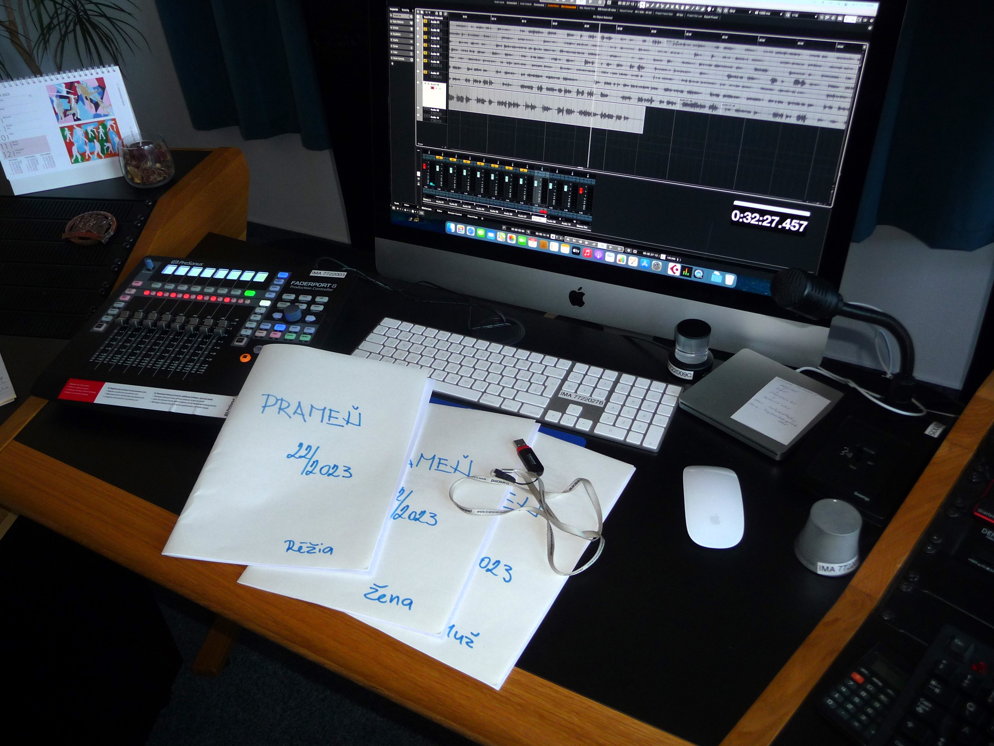 Na obrázku je nahrávací pult zvukového štúdia s textovým podkladom pre réžiu a dvoch spíkrov – muža a ženu.