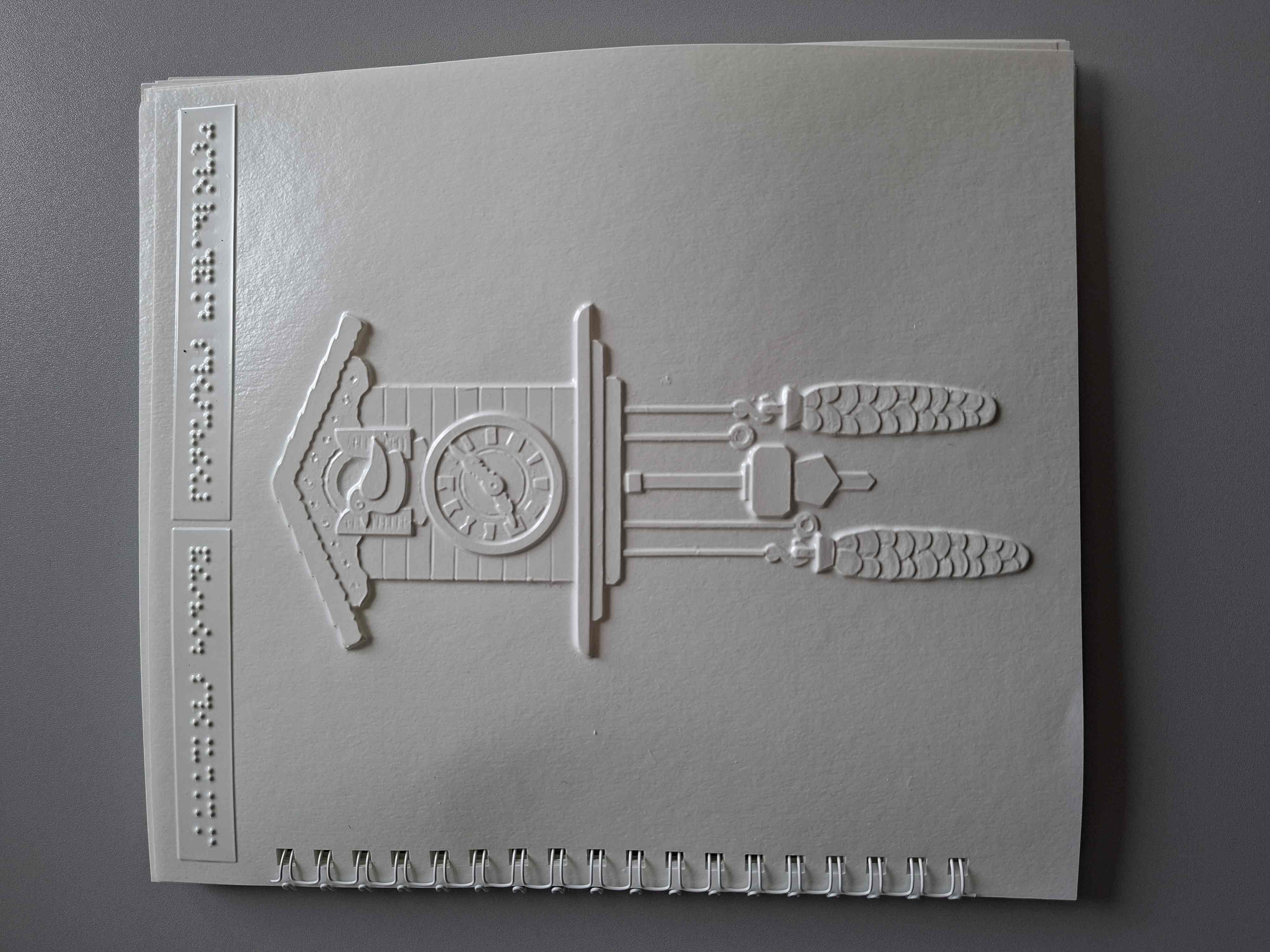 Biely list v špirálovej väzbe s reliéfnym obrázkom kukučkových hodín: hore domček s ciferníkom a malým vtáčikom, dole visia dve šišky na retiazkach a uprostred je kyvadlo.  Hore sú dva malé štítky s braillovým písmom.